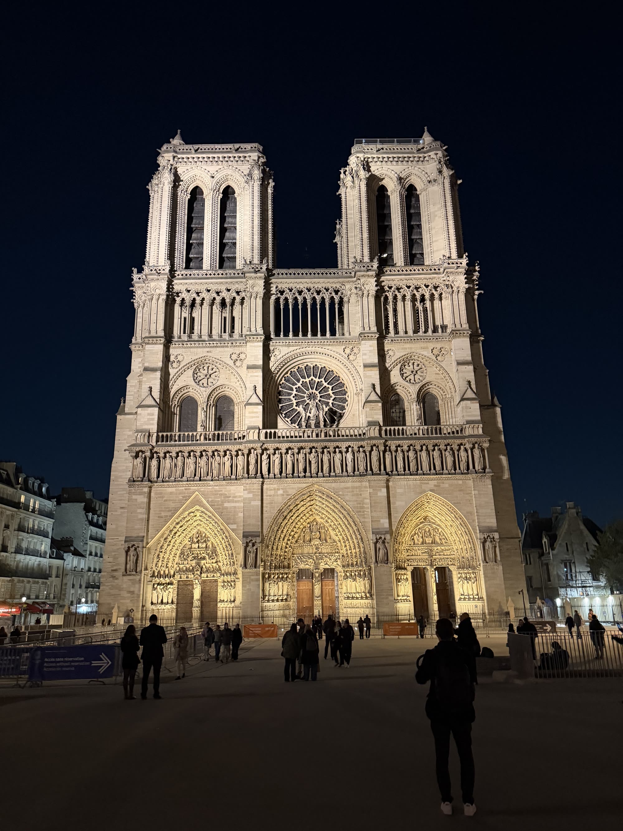 The west front of Notre-Dame lit at night, people crossing the square in front of it