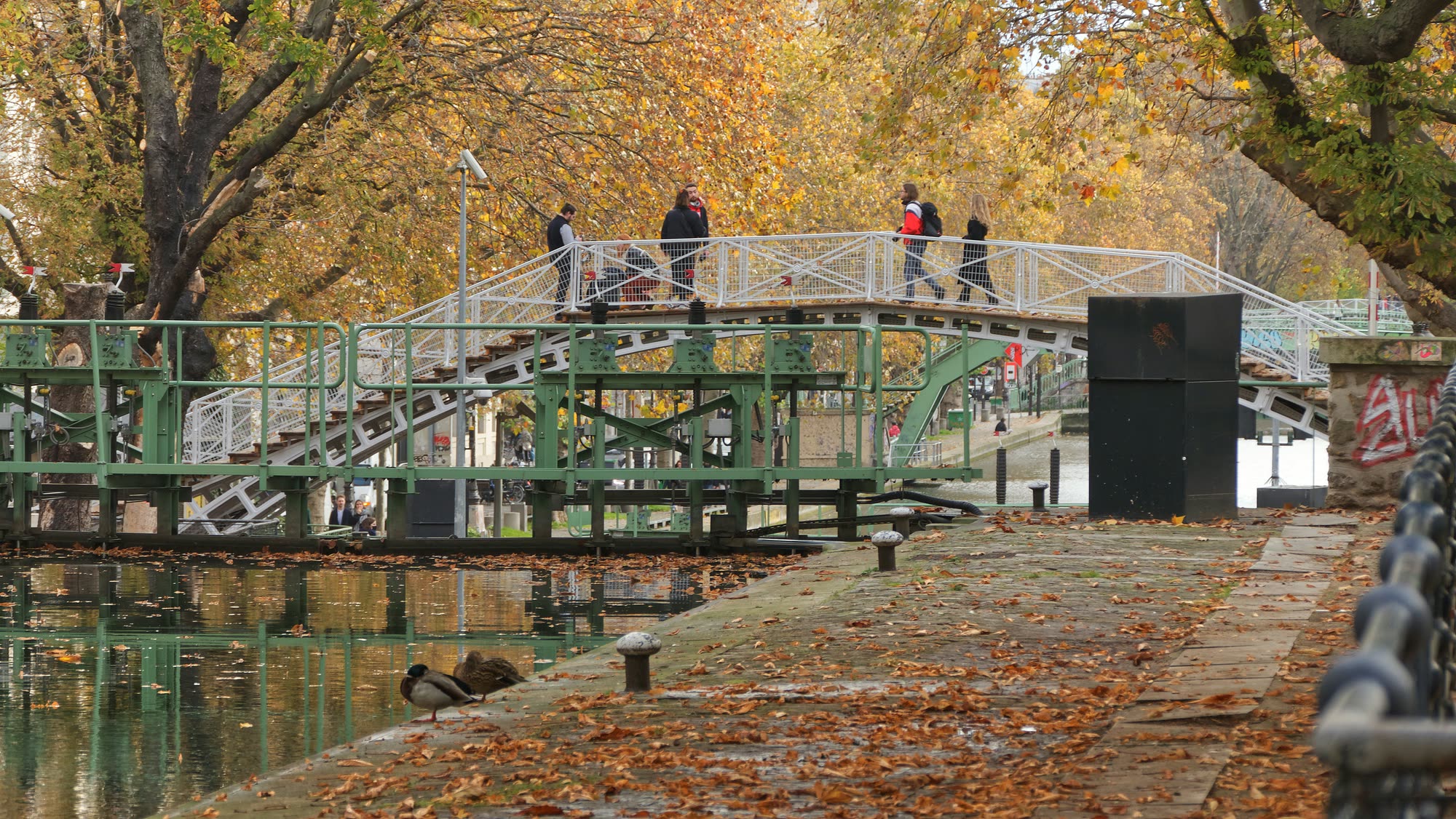 One of the canal's white iron footbridges, with people crossing and leaves on the quay
