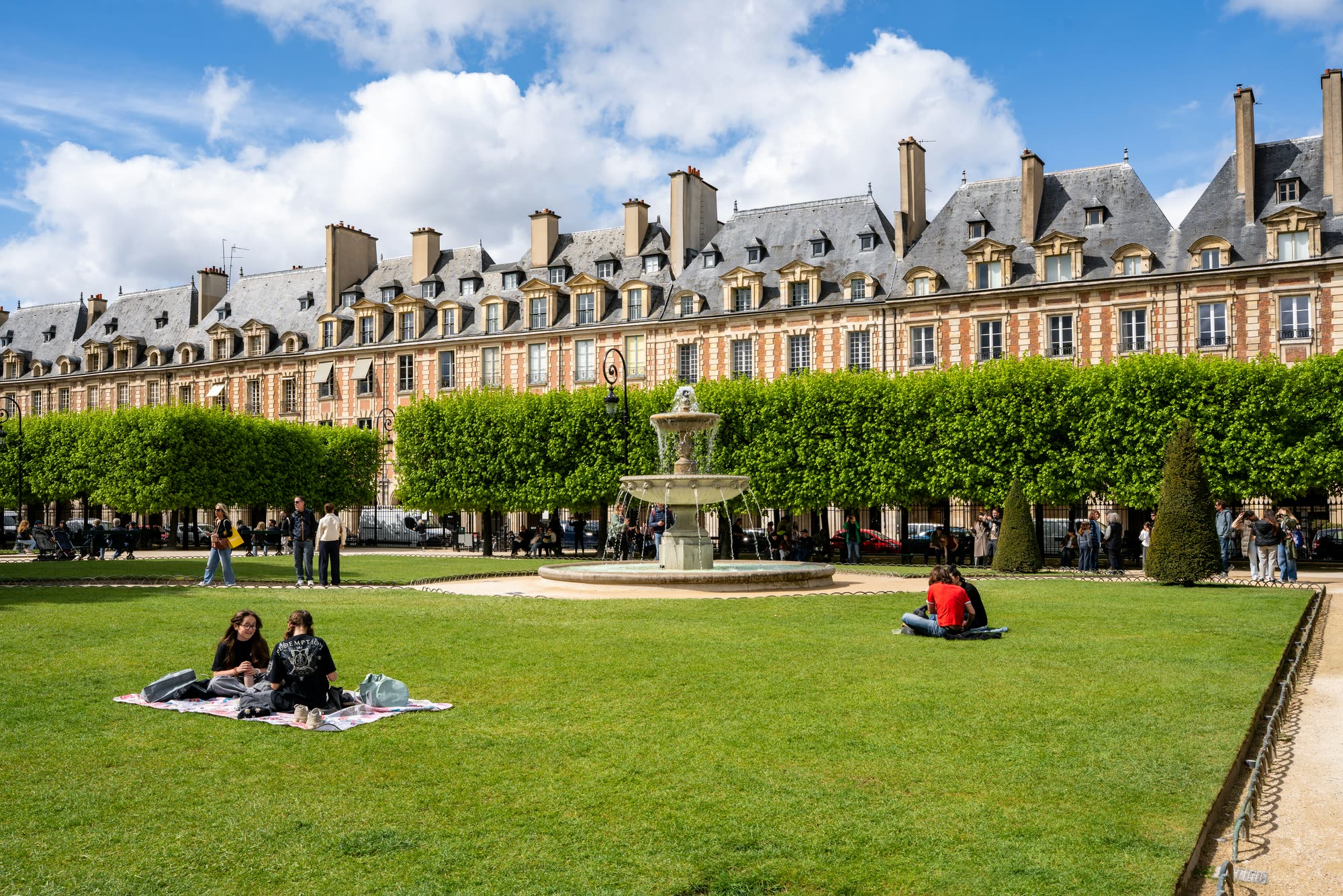 Place des Vosges, red brick façades behind clipped trees, the fountain running and people sitting on the lawn